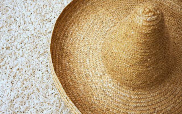 Large sombrero with a twisted border resembling half a torus, made from rows of weaved straws. It's conical tip is blunt and it sits on a white carpet with a fluffy texture.